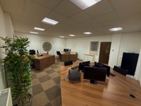 A modern engineering office with several wooden desks, computer monitors, office chairs, a potted plant, a large wall clock, and beige carpet tiles under bright ceiling lights. The room appears clean and organised.