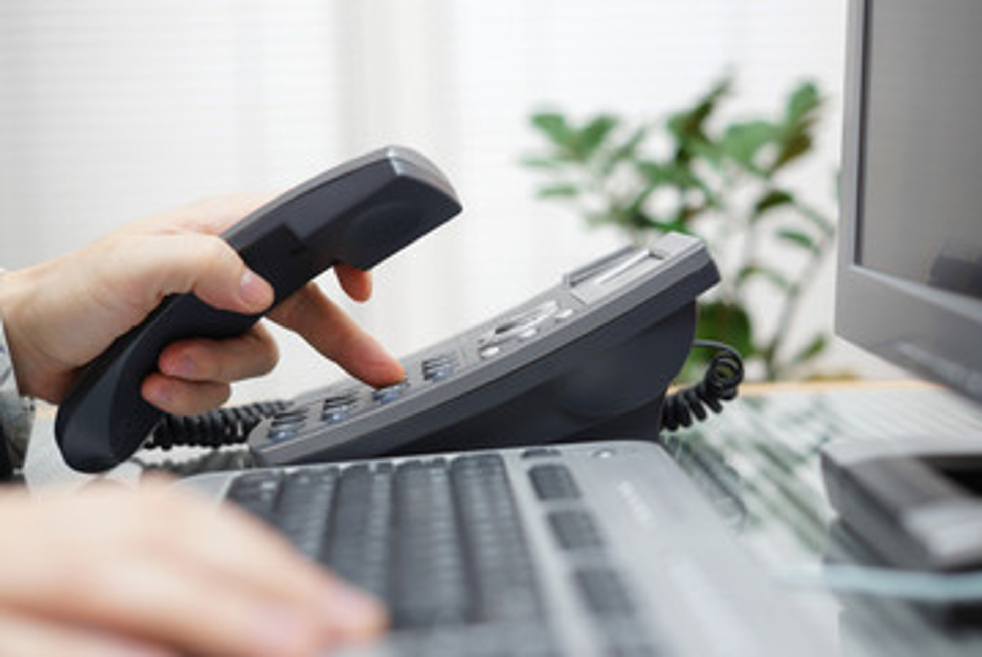 A person dialing a number on a corded office phone while holding the receiver, with a computer keyboard, monitor, and steel machining tools nearby on the desk.