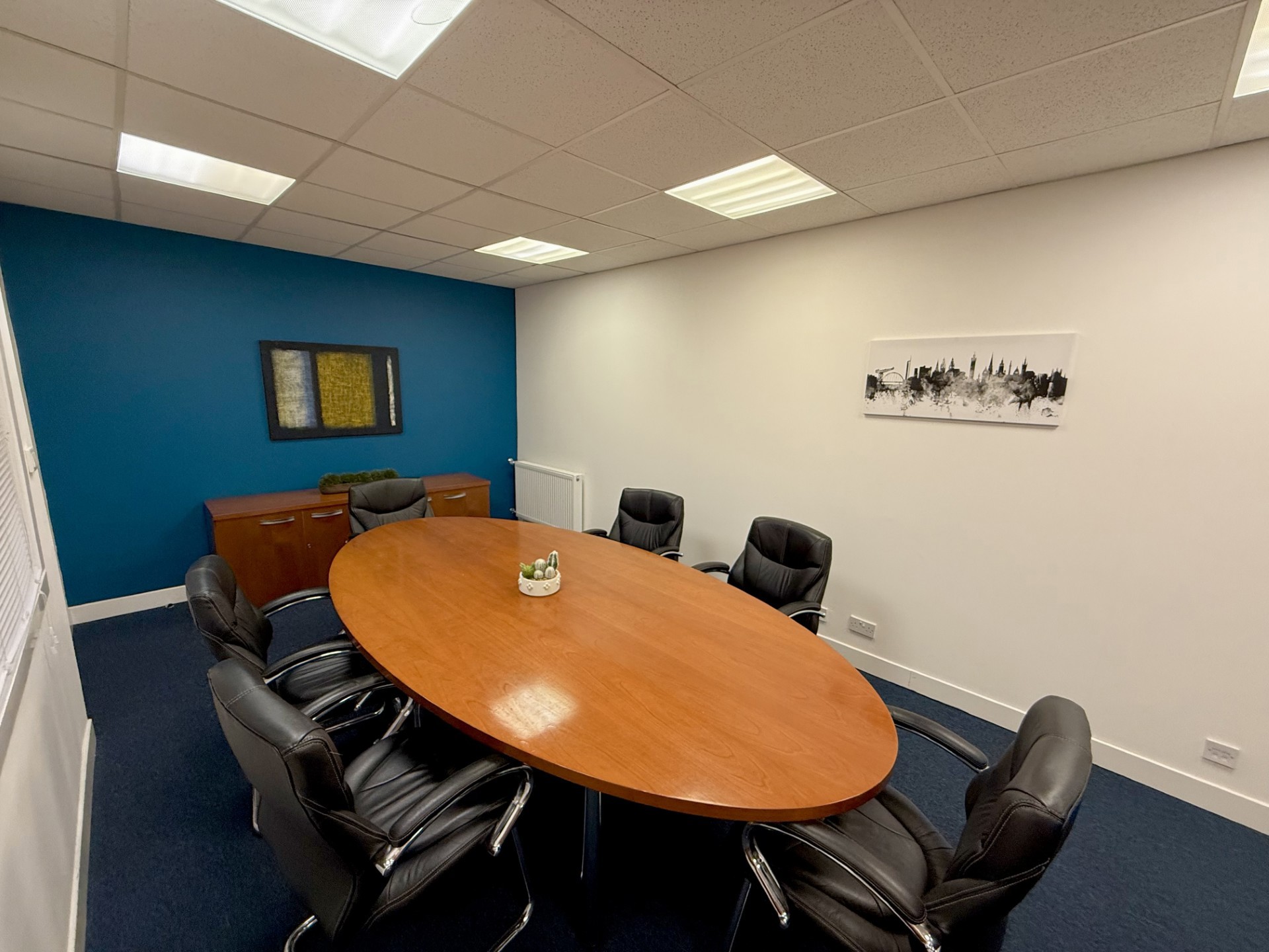 A modern conference room with a large oval wooden table surrounded by eight black leather chairs, blue accent wall with a cabinet, framed picture, and white wall displaying black-and-white artwork of steel engineering.