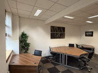 A modern office with four wooden desks arranged in a spacious room, each with a black office chair. The decor features white walls, ceiling lights, and framed artwork, hinting at a sleek manufacturing or steel industry theme. Natural light enters from a window.