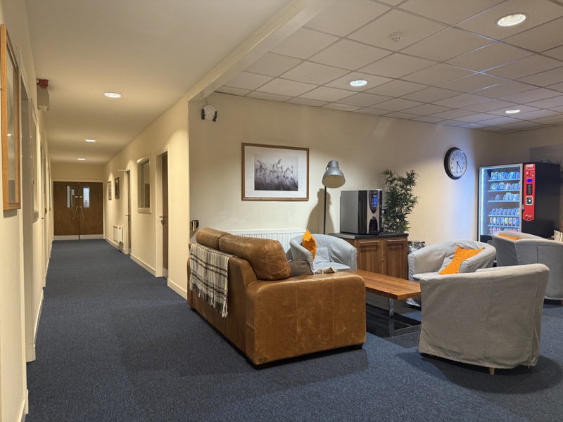 A cozy lounge area with brown and grey sofas, a wooden coffee table, grey carpet, framed picture, clock, vending machine, and a hallway with closed doors and warm lighting, ideal for relaxing in a busy manufacturing or steel stockholder setting.