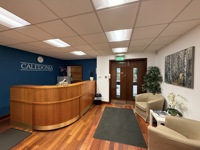 Reception area with a curved wooden desk, blue accent wall with Caledonia sign, two beige chairs, plants, magazine table, and double glass doors. The space reflects Caledonia’s engineering roots with steel accents, wooden floor, and bright ceiling lights.