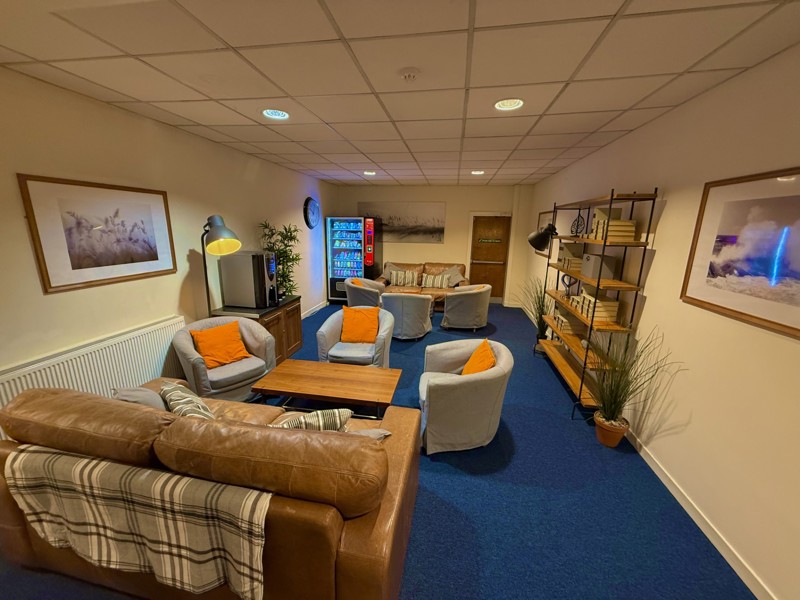 A cozy lounge with blue carpet, brown leather sofa, grey armchairs, orange cushions, a coffee table, bookshelves, wall art, plants, steel accents, vending machine, and a TV on a stand in a warmly lit room.