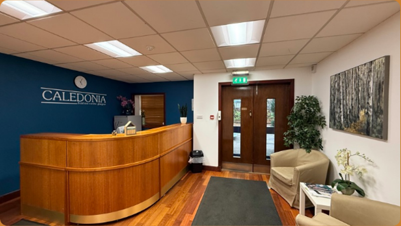 Reception area with a curved wooden desk, two beige chairs, plants, a wall clock, and artwork. The blue wall displays the name Caledonia in steel letters. Large double doors and overhead lights complete the space.