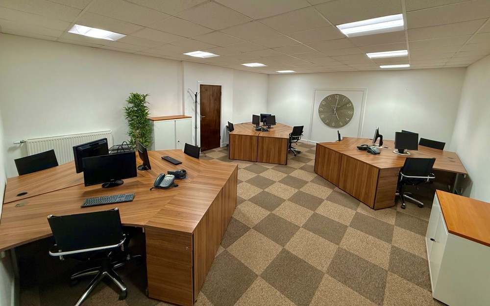 Modern office with six wooden desks arranged around the room, each with a computer and chair, ideal for an engineering or manufacturing team. A large wall clock, potted plant, and neutral-coloured carpeting complete this tidy, well-lit space.