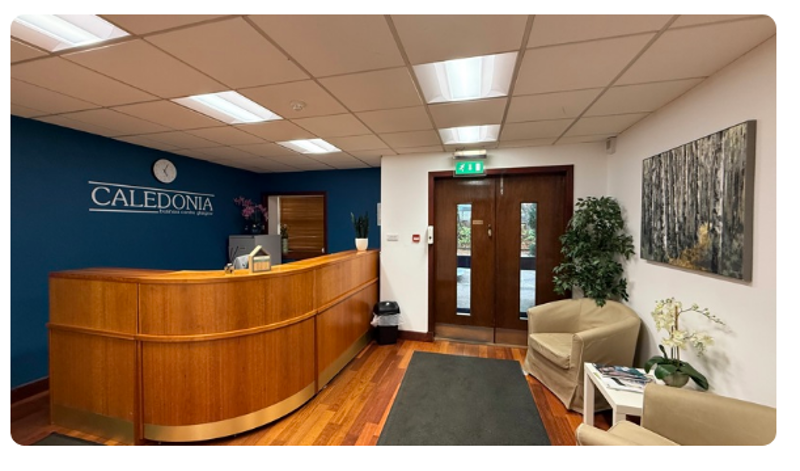 Reception area with a curved wooden desk, blue accent wall with Caledonia sign, armchairs, and a small table with magazines. A painting of trees and a clock reflect the local steel stockholder roots beyond the double glass doors.