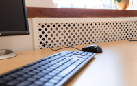A computer keyboard and mouse on a wooden desk, with part of a monitor visible on the left—typical setup found in a serviced office. In the background, beneath a window, a white radiator cover with a lattice design adds to the professional environment often seen at Caledonia Business Centre.