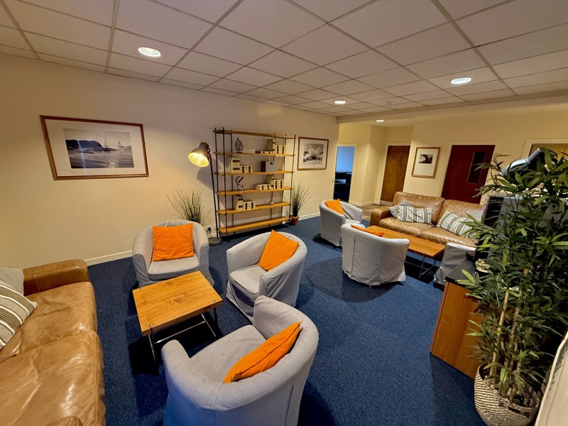A cozy lounge with blue carpet, cushioned chairs and sofas with orange pillows, wooden coffee tables, indoor plants, wall art, and a bookshelf, all under a grid-patterned ceiling with recessed lighting and subtle steel accents.