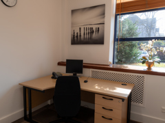 Small office with a desk, black chair, computer, phone, drawers, and a potted plant on the windowsill. A monochrome beach photo hangs on the wall; sunlight streams through the window. Ideal as a serviced office within a business centre, this space offers both functionality and comfort for daily work.