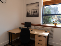 Small office with a desk, black chair, computer, phone, drawers, and a potted plant on the windowsill. A monochrome beach photo hangs on the wall; sunlight streams through the window. Ideal as a serviced office within a business centre, this space offers both functionality and comfort for daily work.