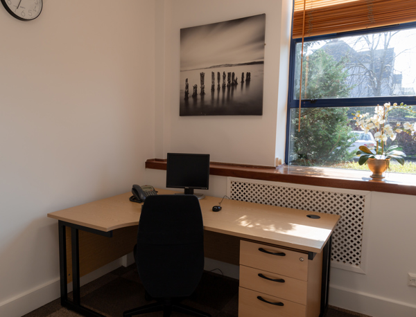Small office with a desk, black chair, computer, phone, drawers, and a potted plant on the windowsill. A monochrome beach photo hangs on the wall; sunlight streams through the window. Ideal as a serviced office within a business centre, this space offers both functionality and comfort for daily work.