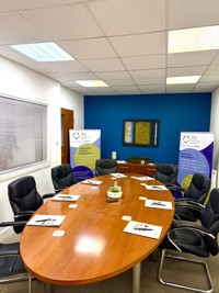A modern conference room with a wooden oval table surrounded by black chairs, meeting materials on the table, and two standing banners by a blue accent wall, ideal for steel stockholder or manufacturing team discussions. A framed artwork and small plant decorate the space.