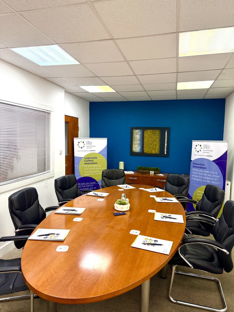 A modern conference room with a wooden oval table surrounded by black chairs, meeting materials on the table, and two standing banners by a blue accent wall, ideal for steel stockholder or manufacturing team discussions. A framed artwork and small plant decorate the space.