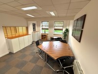 A modern office with geometric wooden desks, black chairs, white storage cabinets, a potted plant, and large windows letting in natural light. Two framed pictures hang on the wall above a carpeted floor in a checker pattern reminiscent of steel machining.