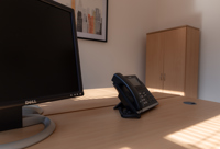 A desk with a Dell monitor, an office phone, and a wooden cabinet in the background, lit by sunlight through window blinds—perfect for a serviced office or business centre environment.