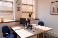 Bright serviced office with two desks, blue chairs, computer monitors, phone, plant, wall art, and a clock. Sunlight streams through wooden blinds on large windows at Caledonia Business Centre in Glasgow.
