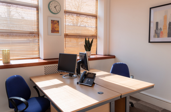 Bright serviced office with two desks, blue chairs, computer monitors, phone, plant, wall art, and a clock. Sunlight streams through wooden blinds on large windows at Caledonia Business Centre in Glasgow.