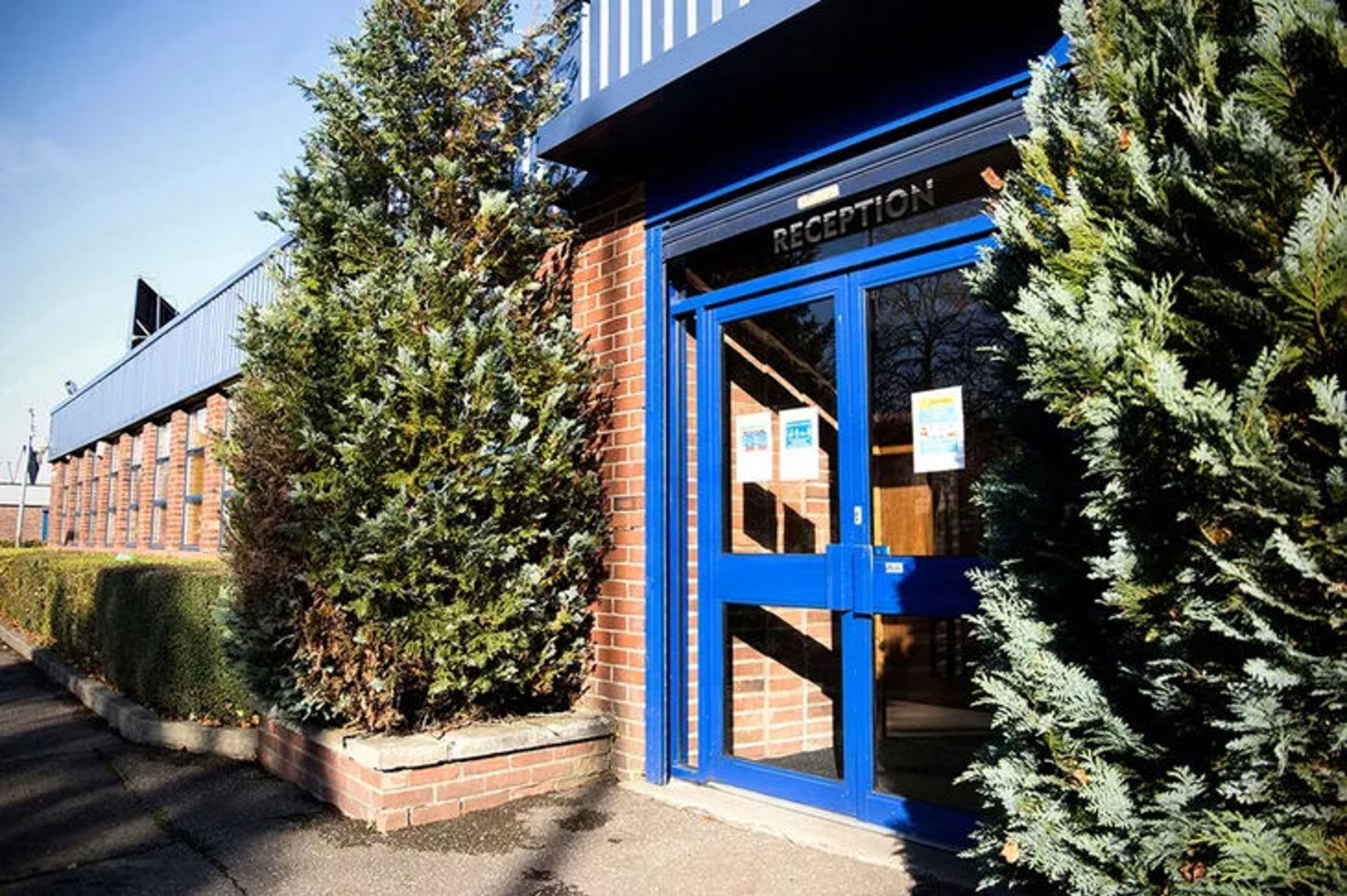 Entrance to a brick building with a blue-framed glass door labeled “Reception,” flanked by two large shrubs; sunlight casts shadows on the path leading to this engineering and manufacturing facility.