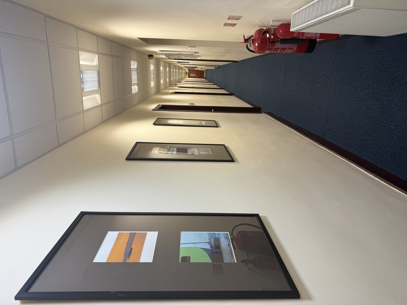 A long hallway with framed pictures on cream-coloured walls, a blue carpet, several doors, ceiling lights, two red fire extinguishers, a white radiator on the right side, and hints of steel accents reminiscent of a manufacturing environment.