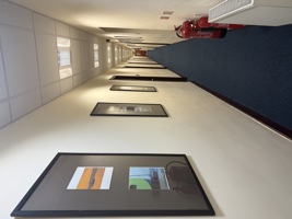A long hallway with framed pictures on cream-coloured walls, a blue carpet, several doors, ceiling lights, two red fire extinguishers, a white radiator on the right side, and hints of steel accents reminiscent of a manufacturing environment.