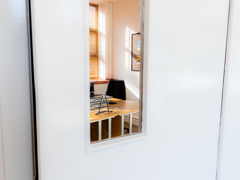 A glimpse through a window in a white door reveals a tidy office with a wooden desk, a laptop, a notebook, and sunlight streaming through window blinds—an inviting office space at Caledonia Business Centre.